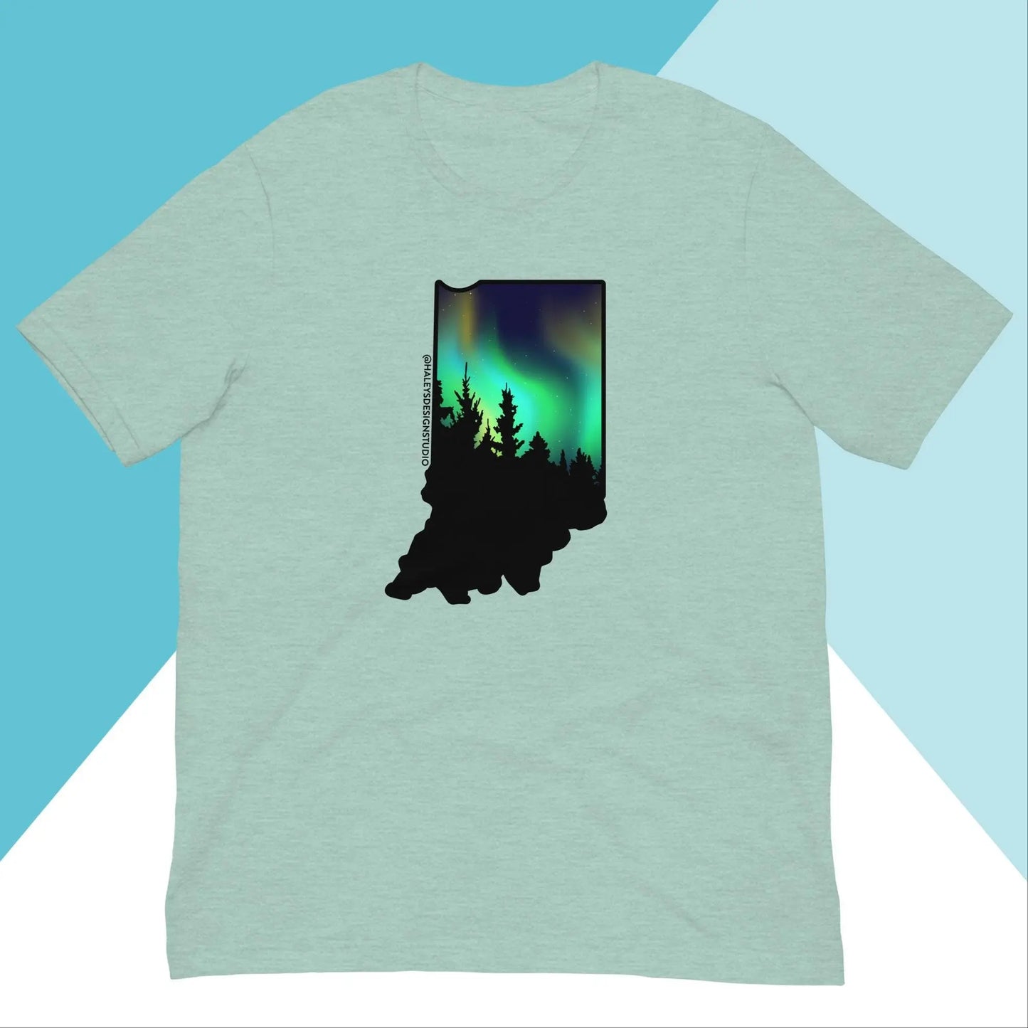 The Indiana Northern Lights Unisex Shirt by North Owl Apparel features a silhouette of Indiana with trees and a northern lights sky. The light green fabric pops against a geometric teal and white backdrop, providing a stylish, comfortable fit.