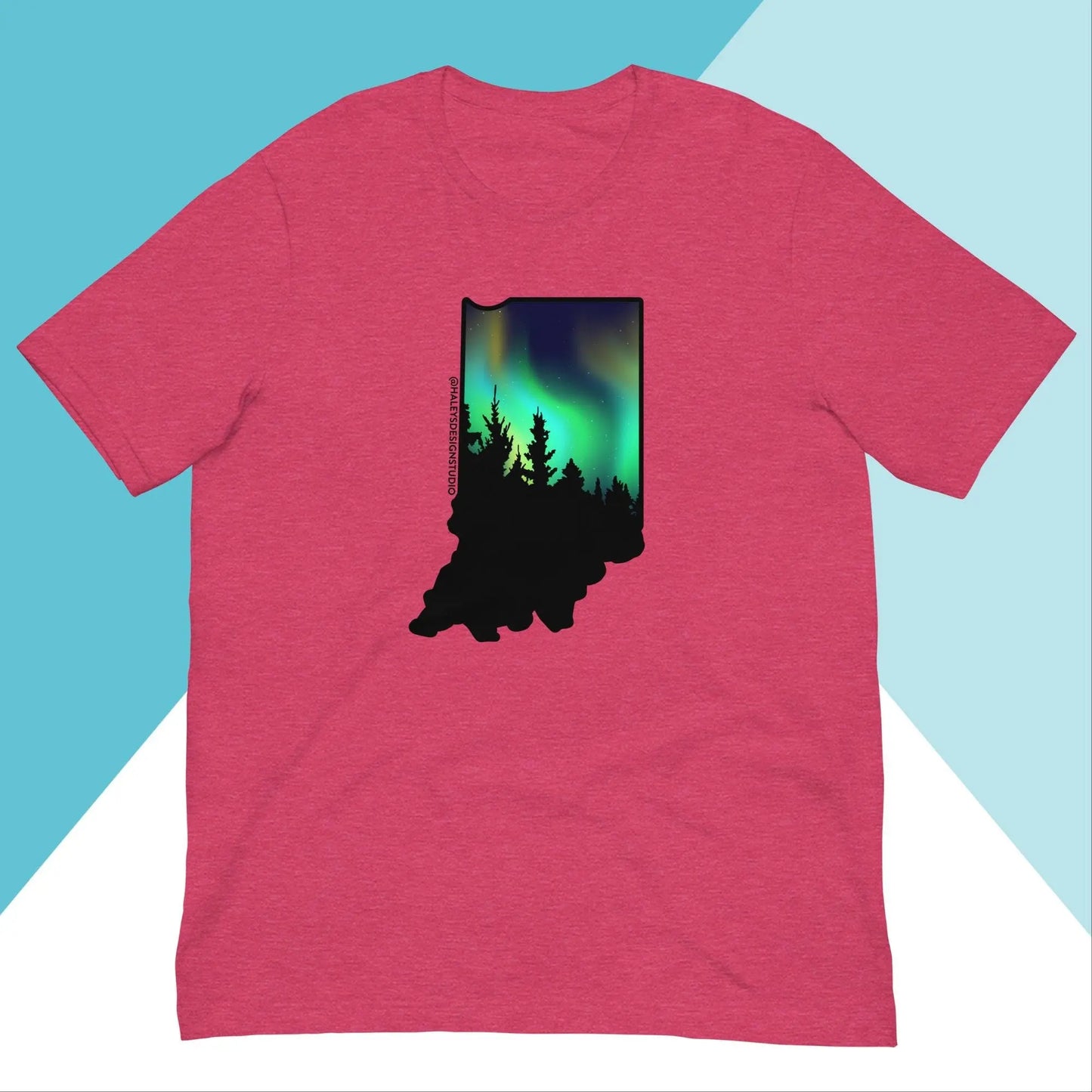 The Indiana Northern Lights Unisex Shirt by North Owl Apparel features a relaxed fit, showcasing tree silhouettes within the Indiana outline. The vibrant greens and blues of the northern lights contrast against a light blue and white geometric background, adding a modern flair.