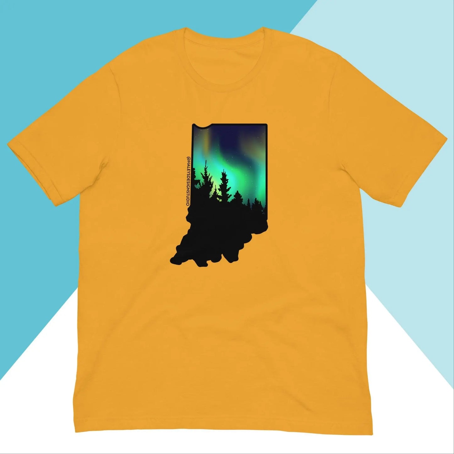 The Indiana Northern Lights Unisex Shirt by North Owl Apparel is a golden-yellow, relaxed fit top featuring a black tree silhouette shaped like Indiana, with green and blue northern lights set against a teal and white diagonal background.