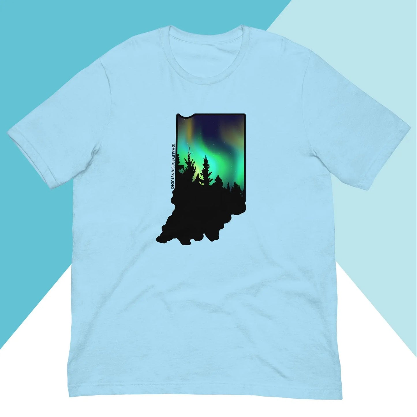 The Indiana Northern Lights Unisex Shirt by North Owl Apparel is a relaxed fit, light blue tee featuring an Indiana silhouette with trees and a vibrant northern lights design on a split light and darker blue background.