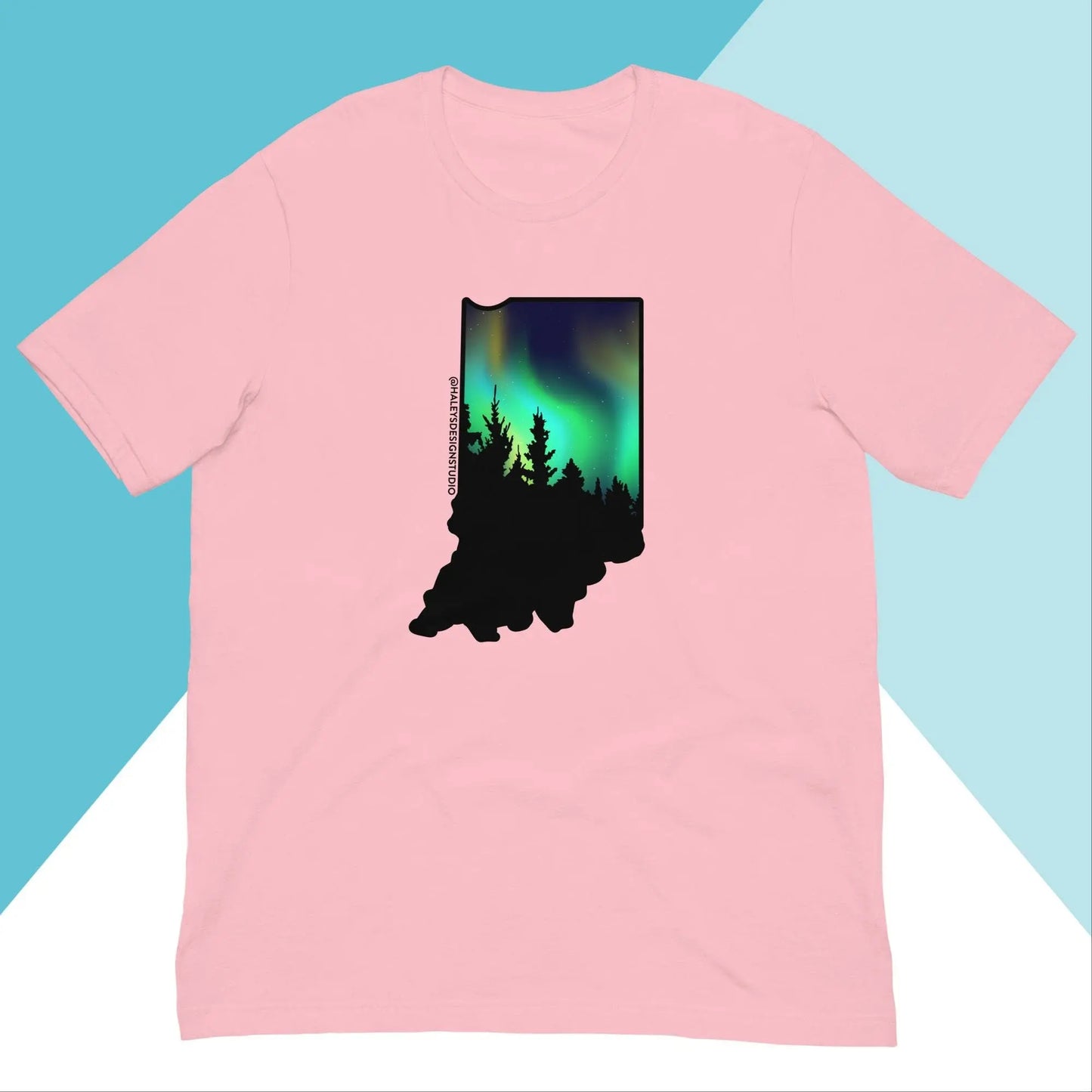 The Indiana Northern Lights Unisex Shirt by North Owl Apparel features a relaxed fit with a pink silhouette of Indiana filled with a forest and northern lights scene against a teal and white geometric background.